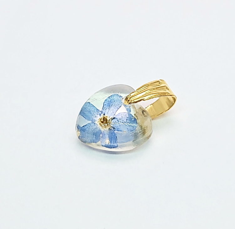 Wisiorek mini serduszko z przejrzystej żywicy, z zatopionym prawdziwym kwiatem niebieskiej niezapominajki. Krawatka w kolorze złotym z pionowymi żłobieniami. Wisiorek ma 1,5 cm długości z krawatką.